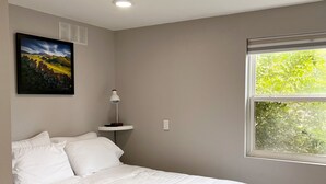 A bedroom with a bed, a window with a view of greenery, a wall-mounted picture, and a ceiling light. - Cozy, modern suite in the heart of Loveland Colorado. (Loveland)