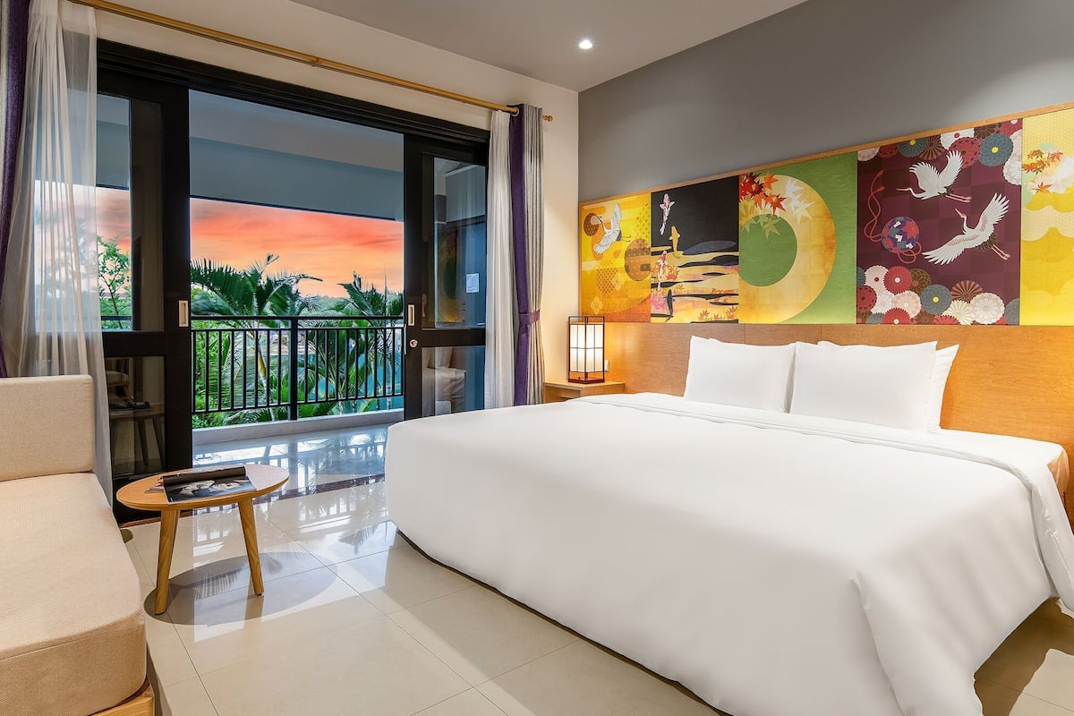 Ein Hotelzimmer mit einem großen Bett, einem kleinen Tisch, einem Nachttisch und Blick auf einen Sonnenuntergang vom Balkon aus.