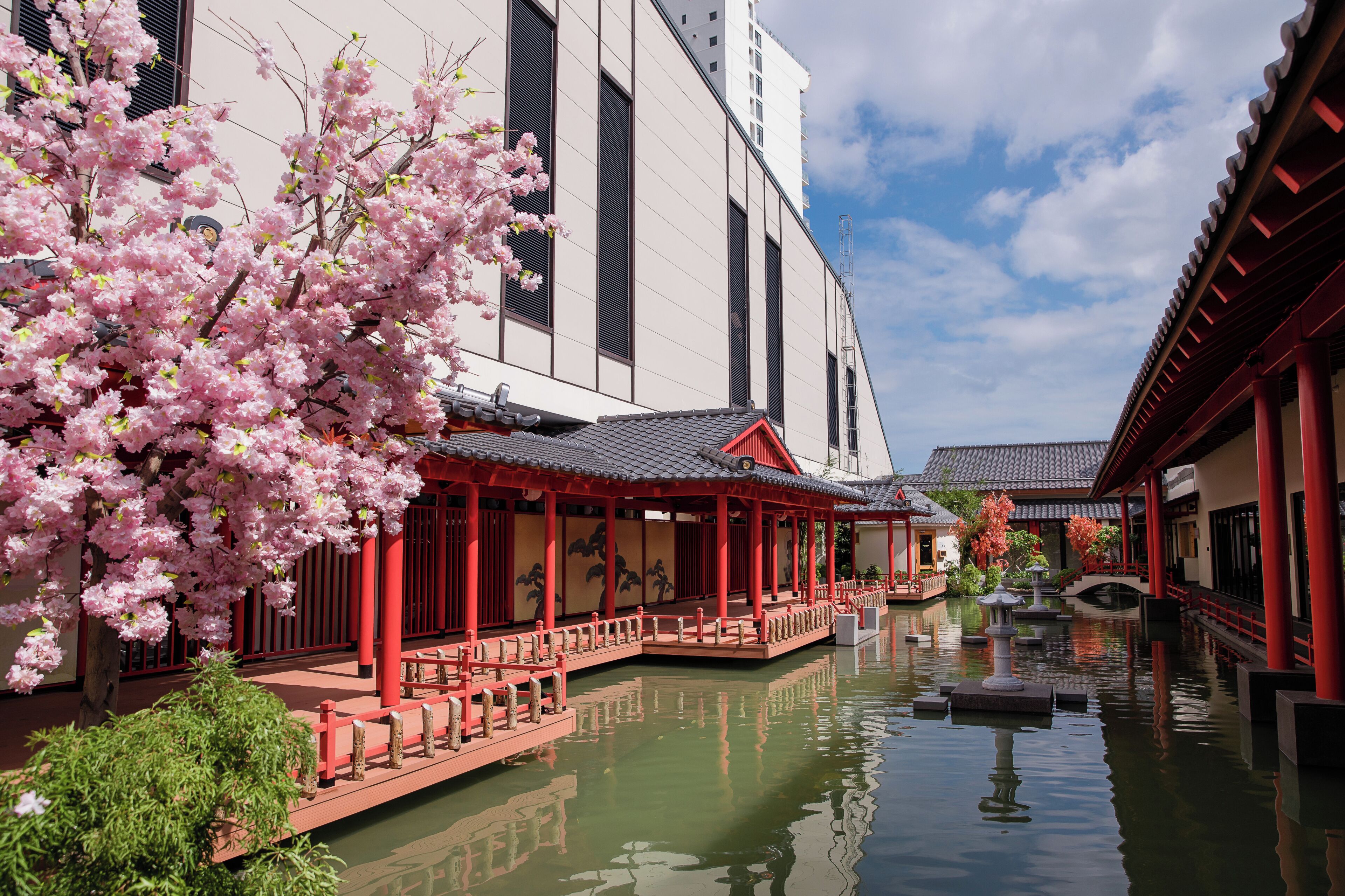 A traditional Japanese building with a water canal, red wooden structures, and cherry blossoms.