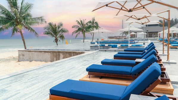 A beachside lounge area with blue cushioned chairs and umbrellas, palm trees, and a view of the ocean. - Mikazuki Villas - The Ryokan (Da Nang)