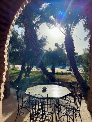 Un espacio de estar al aire libre techado con una mesa y sillas, rodeado de palmeras y vistas a una piscina.