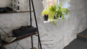 Et værelse med en hvid væg med struktur, et vindue med persienner, en hylde med et kamera og oplader, samt en hængende plante.