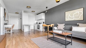 Un salon moderne avec un canapé gris, une table basse et un coin repas, la cuisine étant visible au fond.