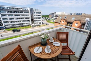 A balcony with a wooden table set for two, a vase with white flowers, and a view of a street with cars and buildings. - Ocean View II - Modern apartment with ocean view, west-facing balcony, Wi-Fi. Pets allowed. (Sylt)