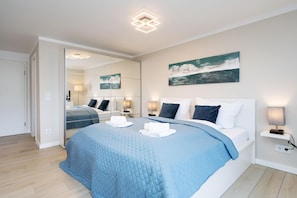 A modern hotel room with a large bed, a mirror, a painting, and a bedside table with a lamp.