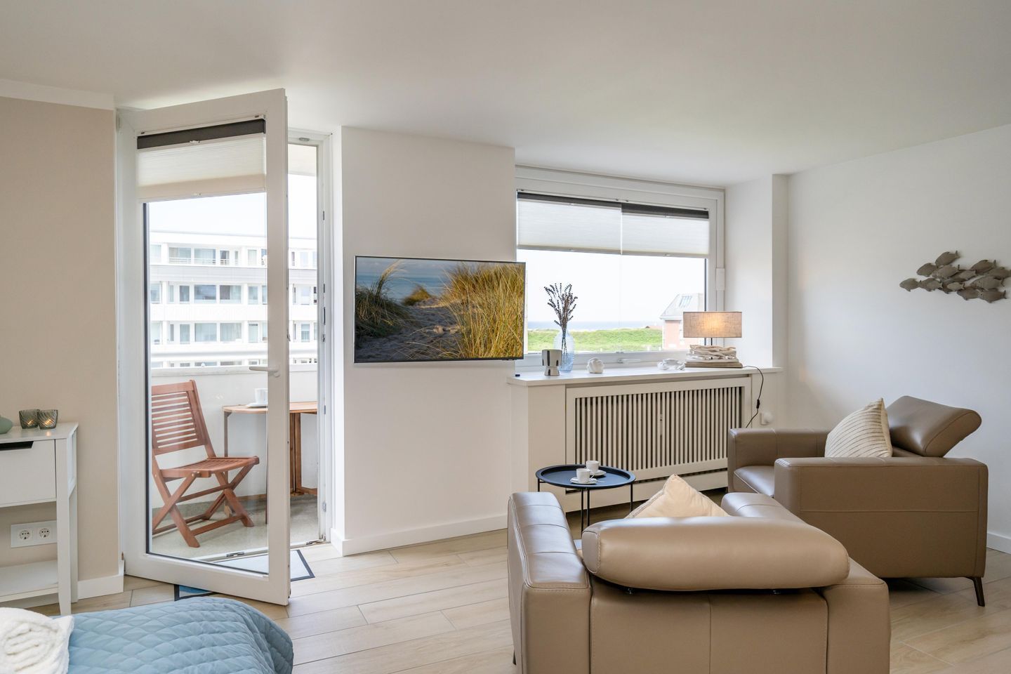 Ein modernes Wohnzimmer mit einer Couch, einem Sessel, einem Couchtisch und einem Fernseher auf einem Standfuß. Ein Fenster bietet Blick auf Gebäude, und eine Tür führt in einen Außenbereich.