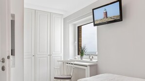 A bedroom with a bed, a small table with a chair, a radiator, a window with a view of a lighthouse, and a TV mounted on the wall. - SyltMomente - 2-room apartment close to the beach: balcony, parking space, WLAN. Pets (Sylt)
