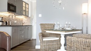 A modern kitchen with grey cabinets, a built-in microwave, and a coffee machine. There is a dining area with wicker chairs and a glass table set for four. - SyltMomente - 2-room apartment close to the beach: balcony, parking space, WLAN. Pets (Sylt)