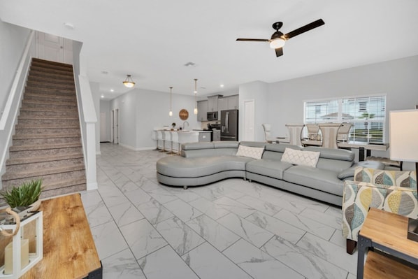 A spacious living area with a curved gray sectional sofa, a wooden coffee table, and a ceiling fan. A kitchen with modern appliances is visible in the background.