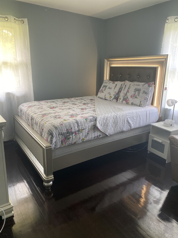 A neatly made bed with floral bedding and a headboard with illuminated panels, a bedside table with a lamp, and a dark wooden floor. - Akeem's  guest house (Silver Spring)