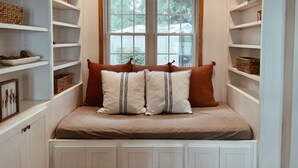A cozy reading nook with a cushioned bench, a window with a view of trees, and shelves with baskets and books. - Maple Haven :: Modern, year round home on Maple Lake in Mentor, MN (Mentor)