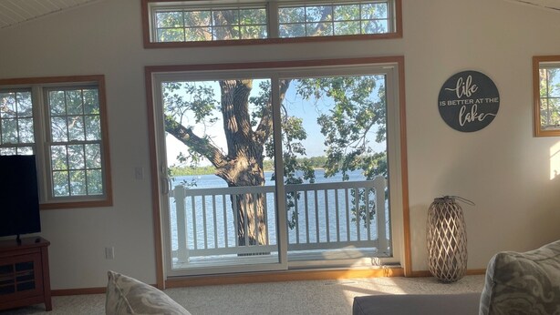 A living room with a large sliding glass door leading to a deck overlooking a lake, a television on a stand, and a decorative wall sign. - Maple Haven :: Modern, year round home on Maple Lake in Mentor, MN (Mentor)