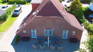 Ein zweistöckiges Backsteingebäude mit geneigtem Dach, einem zentralen Eingang und einer Terrasse mit Gartenmöbeln.