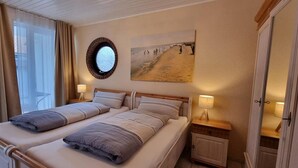 Habitación de hotel con dos camas, un espejo redondo, un cuadro de playa y una ventana con cortinas.