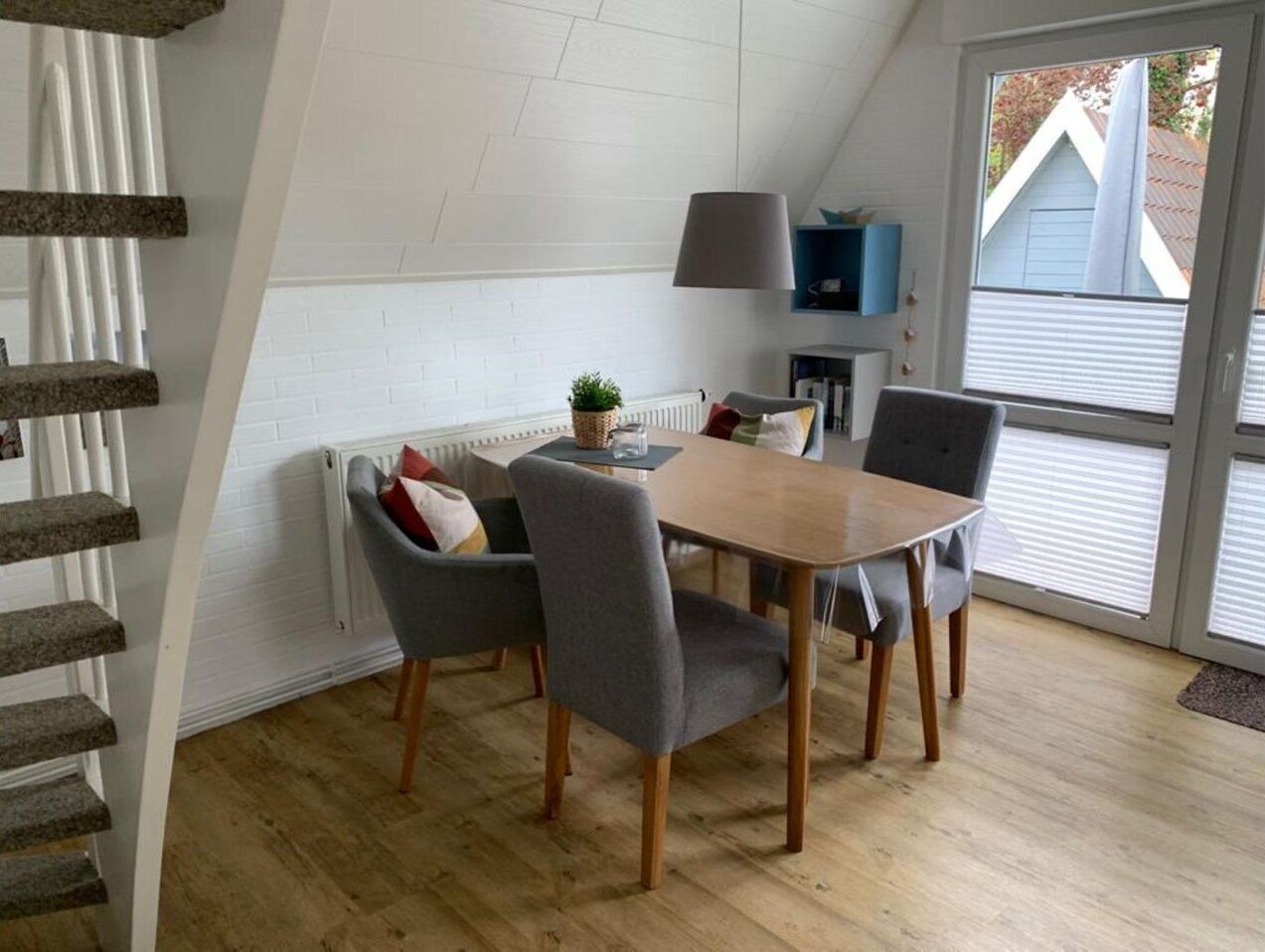 A dining area with a wooden table and four grey chairs, a small plant on the table, and a grey lamp on the wall.