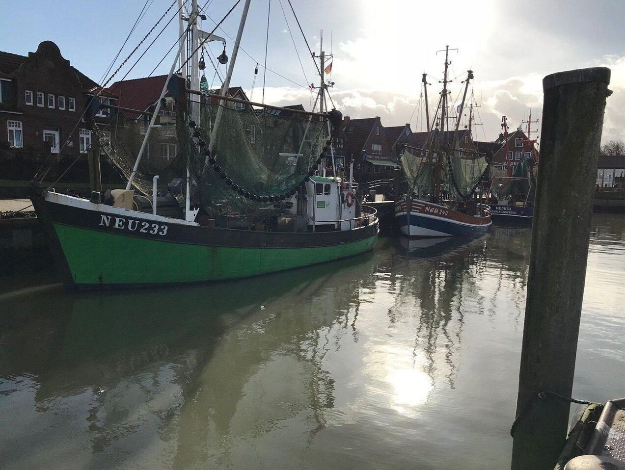 A harbor with fishing boats docked, a wooden pier, and buildings in the background.