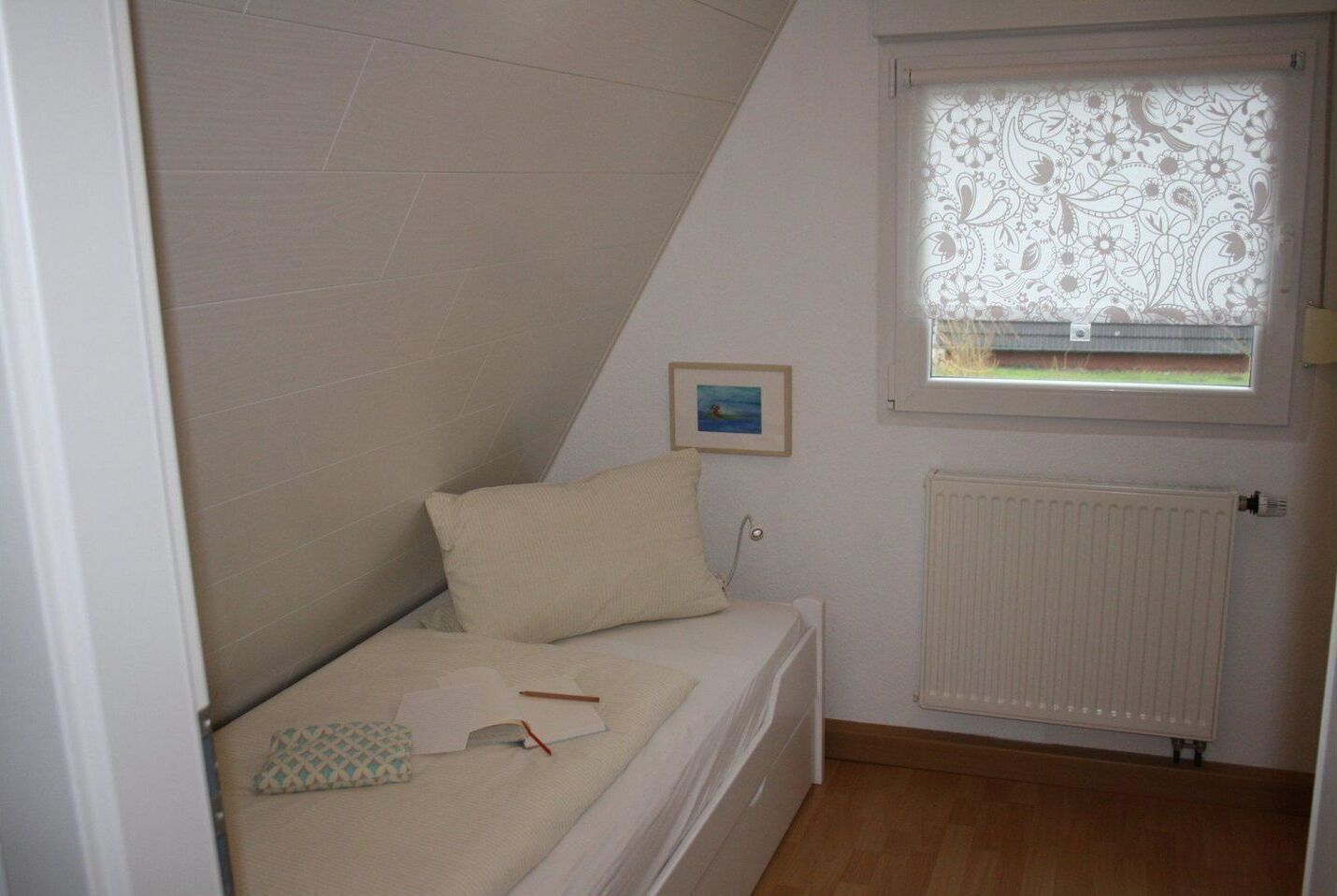 Une petite chambre avec un lit simple, un plafond en pente, un radiateur, une fenêtre ornée d’un motif floral et un tableau encadré accroché au mur.