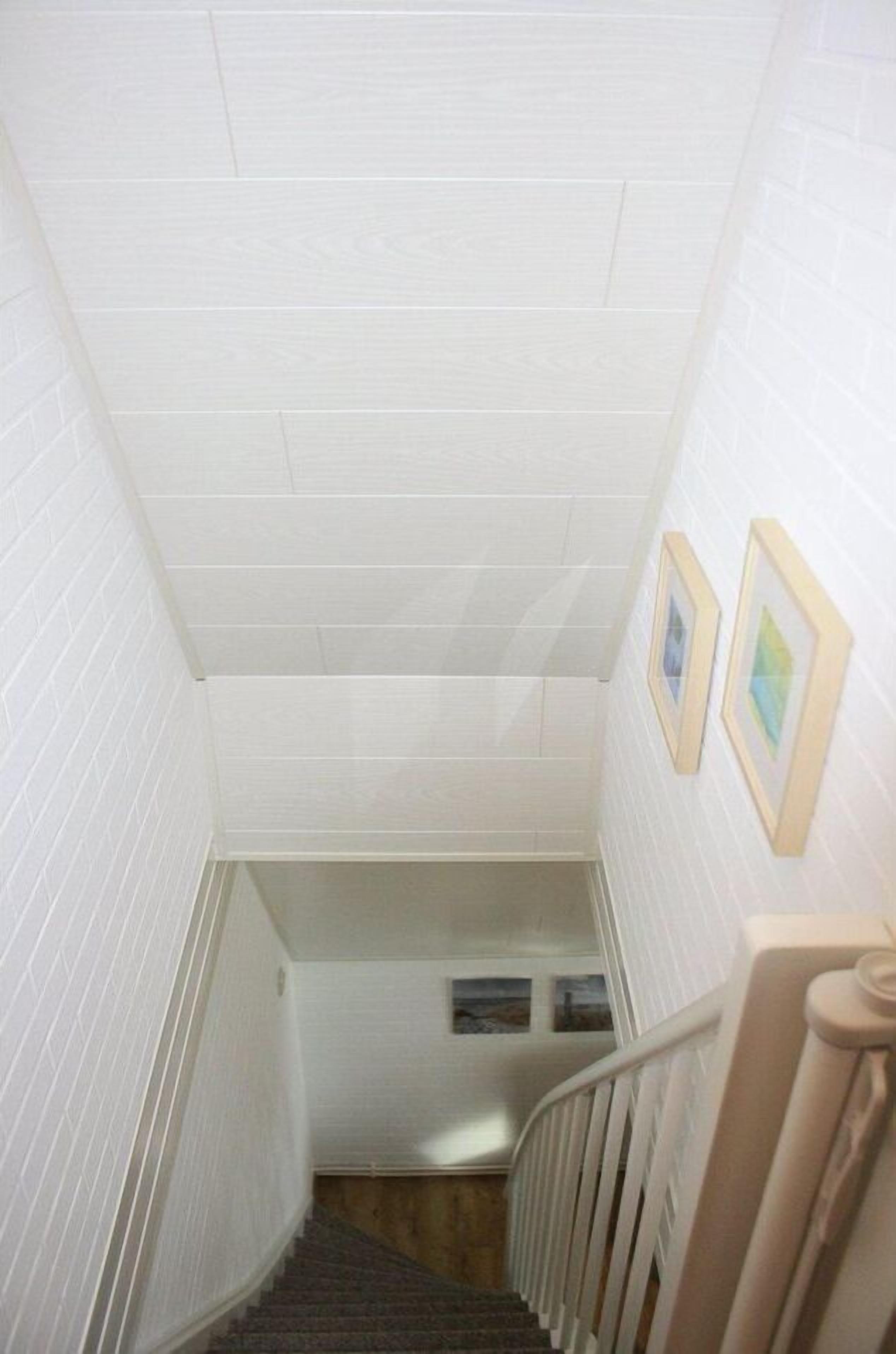 Un escalier étroit aux murs blancs et au plancher en bois, menant à un palier doté d’une rambarde blanche.
