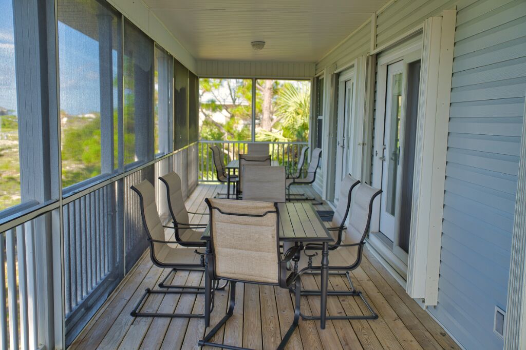A covered porch with a table and chairs, large windows, and a view of greenery.