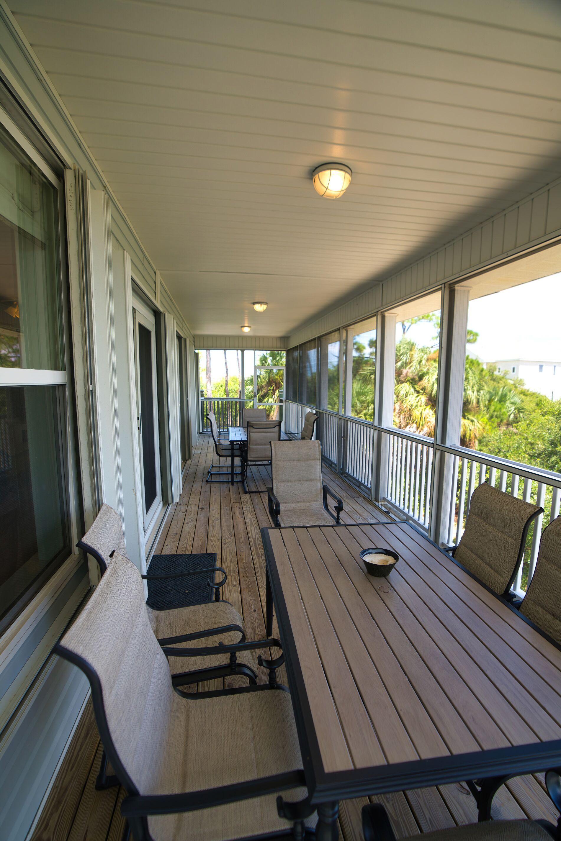 A covered outdoor area with a wooden table and chairs, large windows, and a view of greenery.