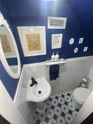 Une salle de bain avec un lavabo blanc, des toilettes et un miroir. Des tableaux encadrés ornent le mur, et une étagère contient des articles de toilette.