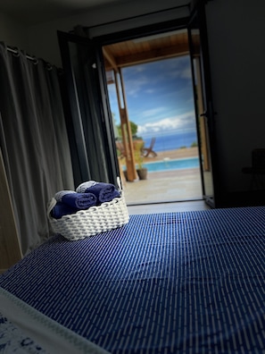 Une chambre avec un lit, un panier en osier contenant des serviettes, et une vue sur la piscine et la plage, visible par une porte ouverte.
