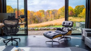 A modern office with a large desk, office chair, and a view of autumn trees through large windows. - Hudson Ridge / Hot Tub, Fire Pit & Fireplace (Hudson)