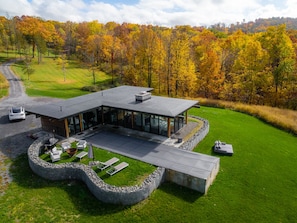A modern house with a large glass facade, surrounded by a stone retaining wall, set in a lush green landscape with autumn-colored trees.