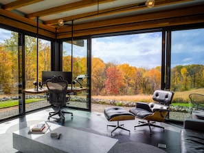 A modern office with large windows, a desk, office chair, and leather ottomans.