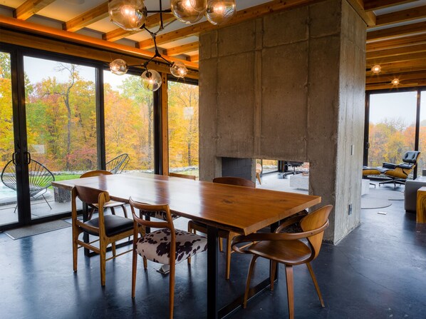A modern dining area with a large wooden table and chairs, a concrete accent wall, and large windows offering a view of autumn trees.