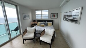 Een slaapkamer met een bed, twee stoelen, een nachtkastje, een raam met uitzicht op de oceaan en kunst aan de muren.