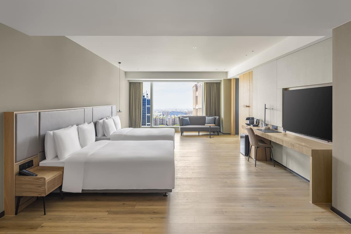 A modern hotel room with a large bed, a desk, a TV, and a view of the cityscape.