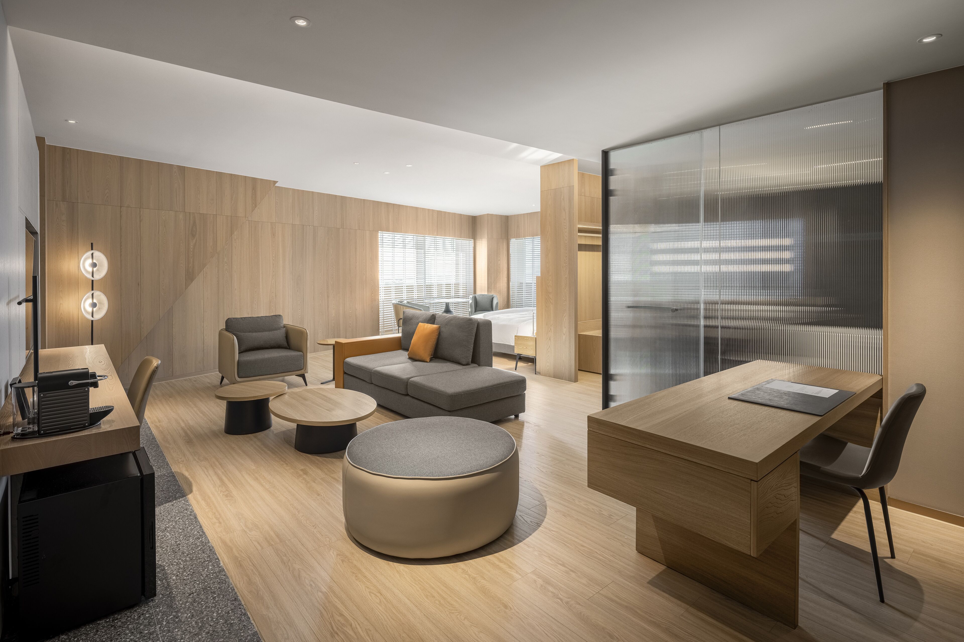 A modern hotel room with a wooden floor, a sofa, a round ottoman, a desk with a chair, and a large window with blinds.