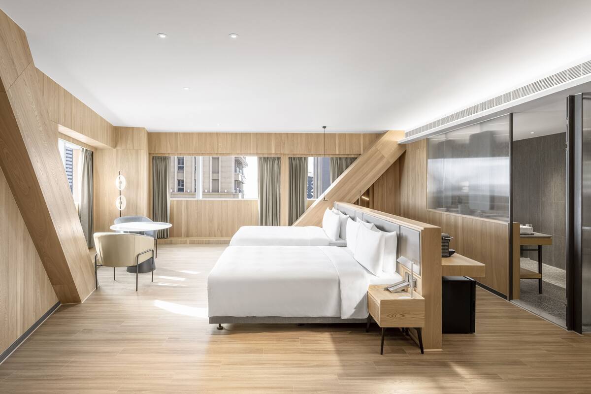 A modern hotel room with a large bed, a desk, and a view of the city through large windows.