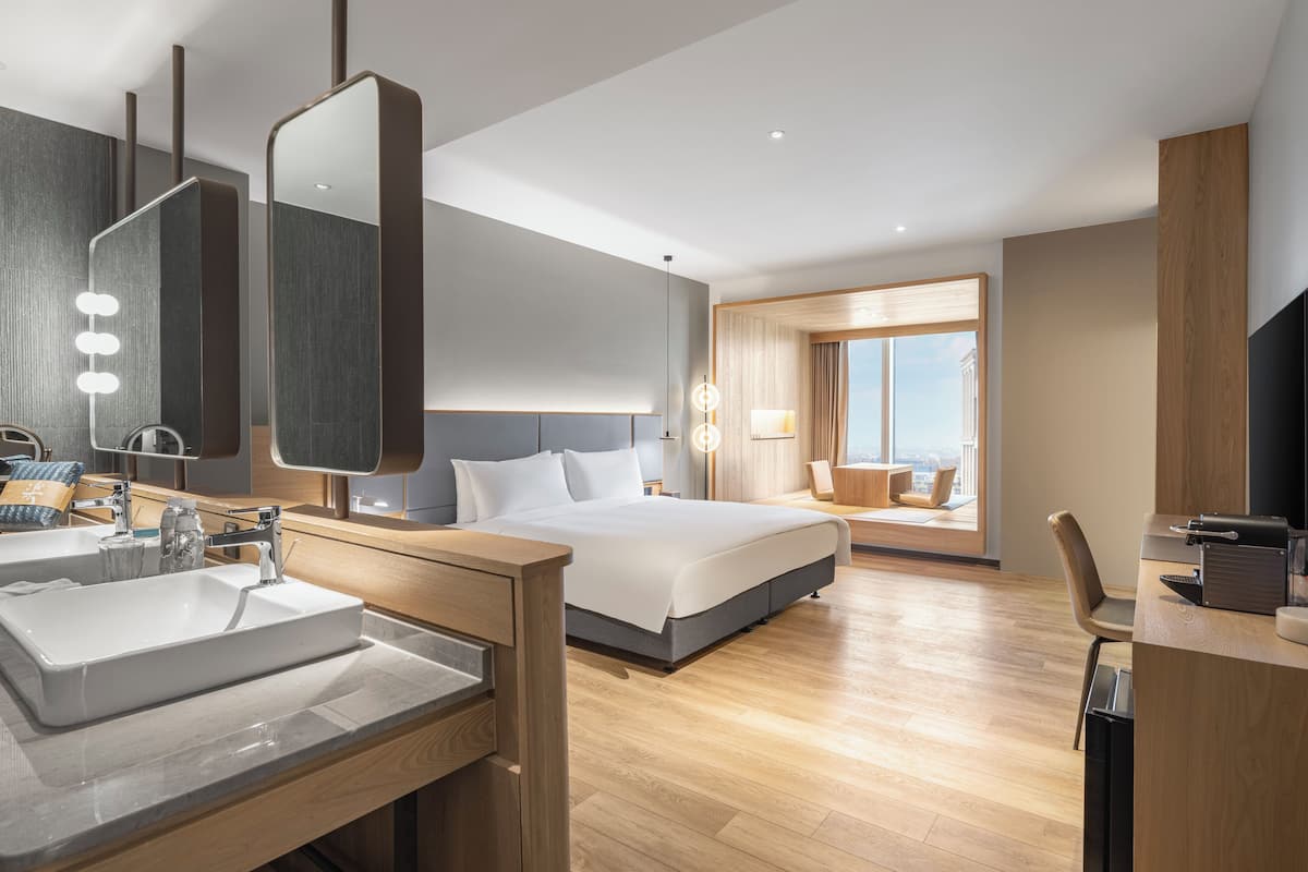 A modern hotel room with a large bed, a desk with a computer, a bathroom with a sink and mirror, and a large window offering a city view.