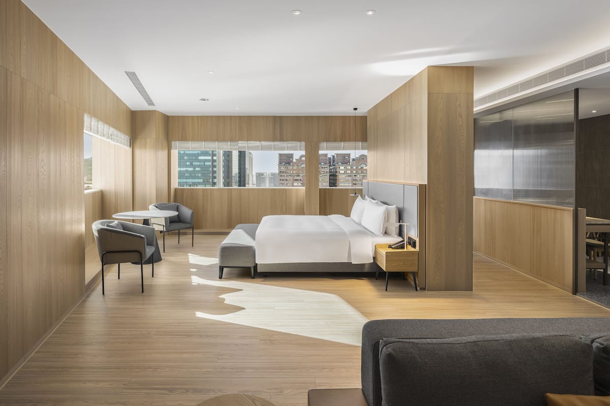 A modern hotel room with a large bed, a sitting area with a table and chairs, and a city view through large windows.
