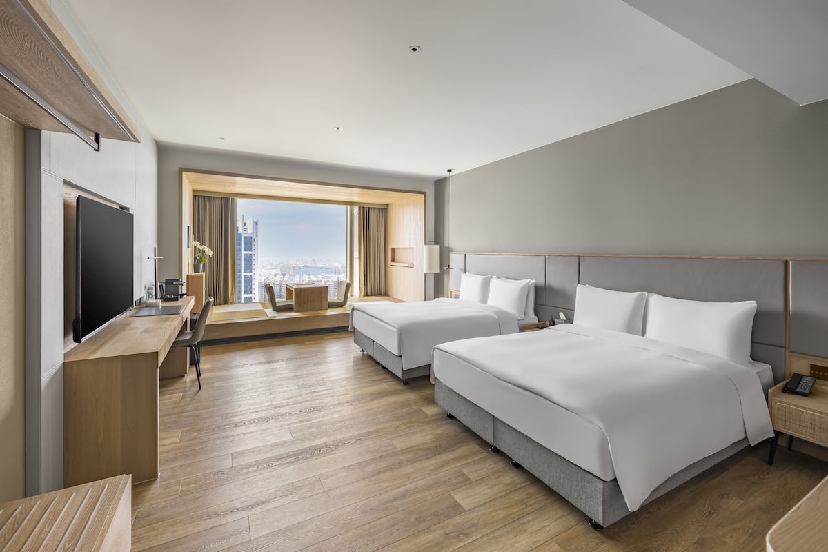 A modern hotel room with two beds, a desk, a television, and a large window offering a city view.