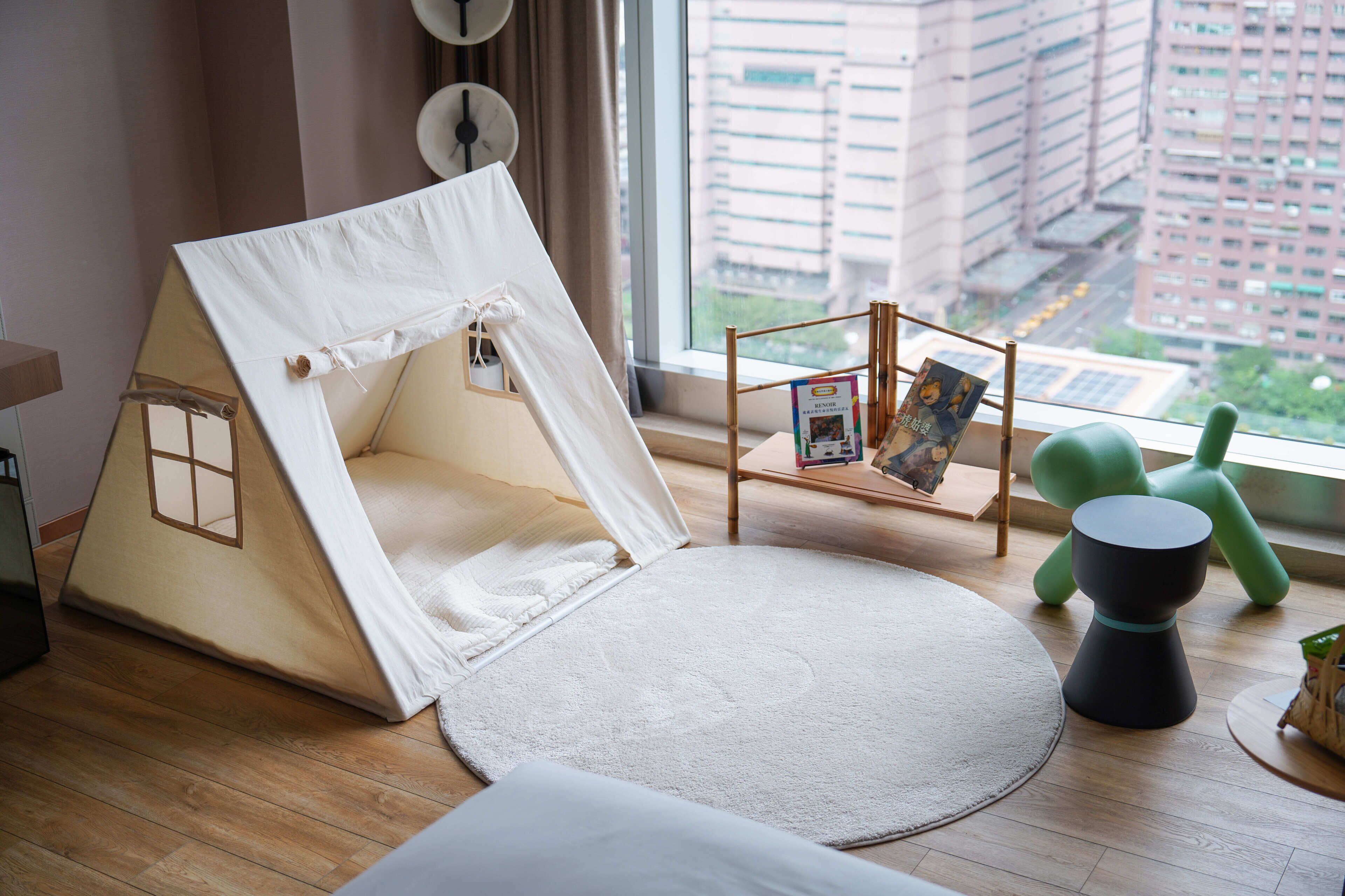 A room with a large window, a white tent-like structure, a round rug, a small table with a book, and a green chair.