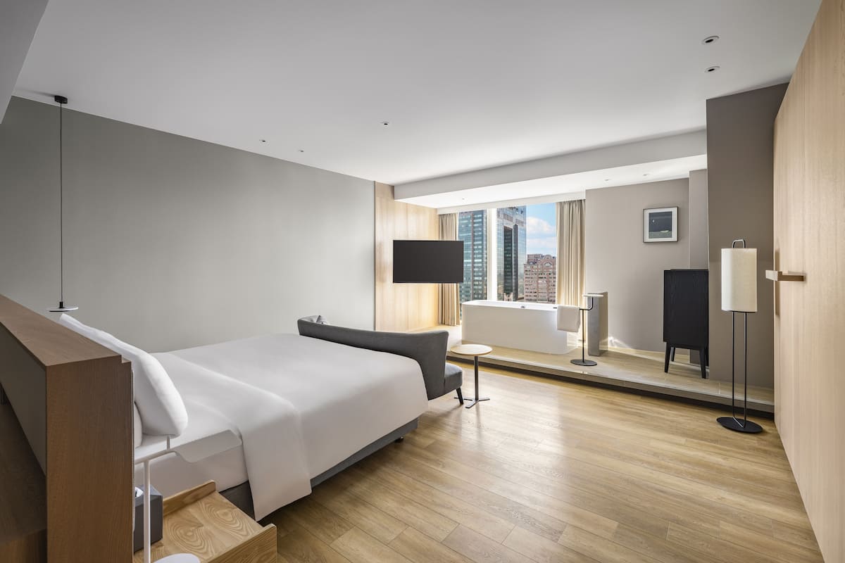 A modern hotel room with a large bed, a small sofa, a desk, and a large window offering a city view.