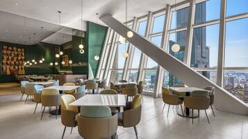 A modern dining area with tables and chairs, large windows with a city view, and hanging spherical lights.