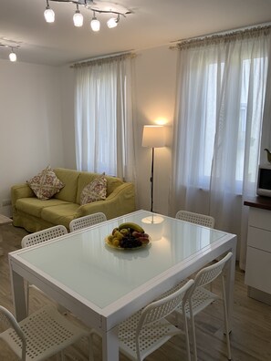 A dining area with a glass table, white chairs, a green sofa with patterned cushions, and a lamp.