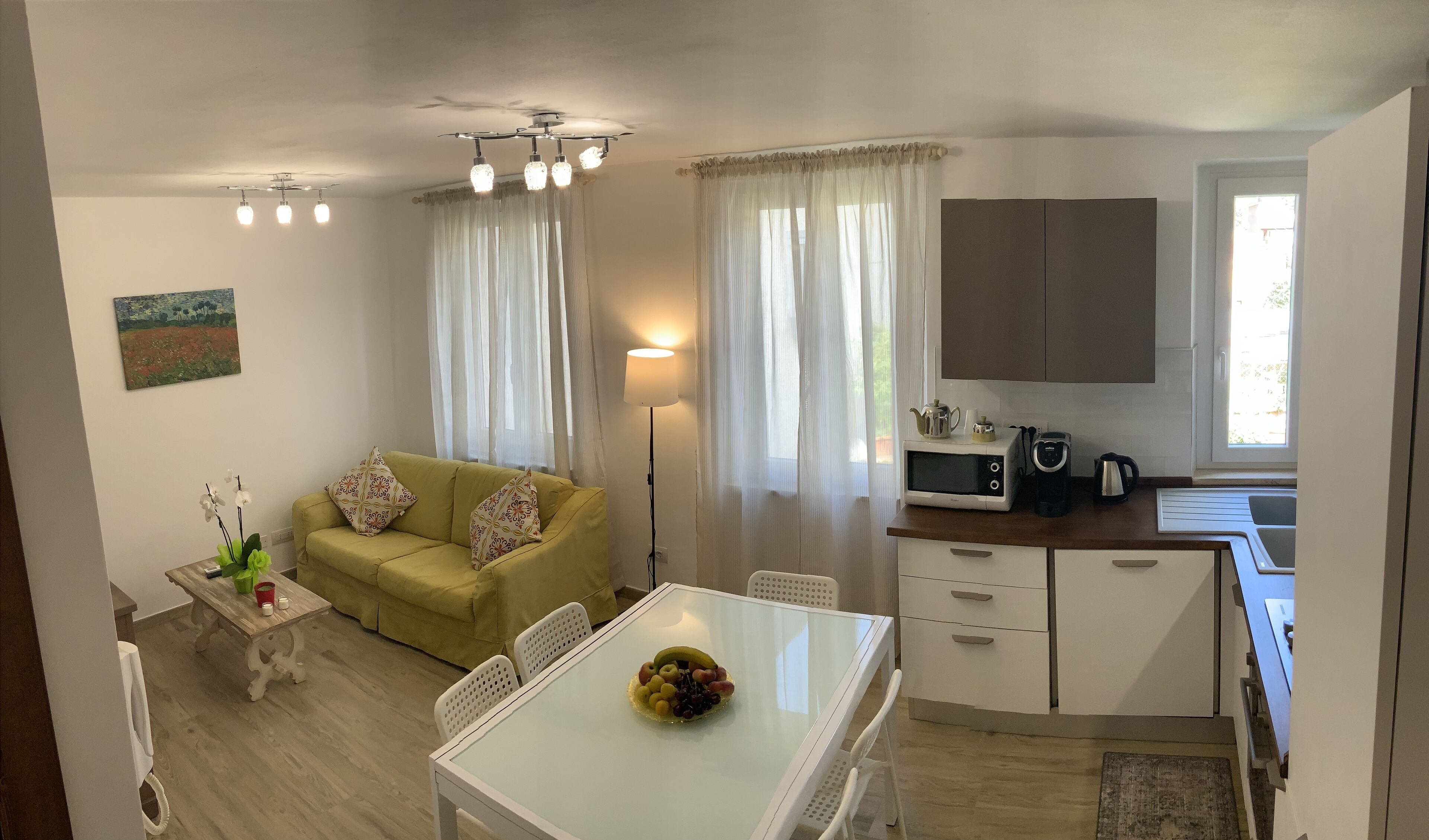A compact living space with a sofa, a dining table with fruit, a kitchen area with appliances, and sheer curtains.