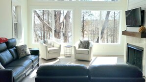 A spacious living room with a large window overlooking a forest, a fireplace, and a television mounted on the wall.
