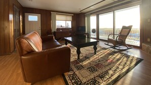 Ein geräumiges Wohnzimmer mit einem braunen Ledersofa, einem dunkelholzigen Couchtisch und einem gemusterten Teppich. Ein Fernseher steht auf einem Schrank, ein Schaukelstuhl und ein großes Fenster mit Blick auf eine Holzterrasse.