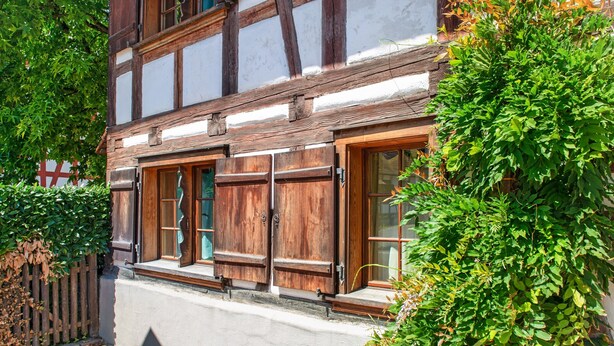 Un bâtiment traditionnel à colombages, doté de volets en bois, d’un chemin pavé et d’une végétation luxuriante.