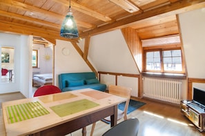 A cozy room with a wooden ceiling, a blue sofa, a wooden table with placemats, a red chair, a television, and a window with blinds.