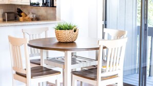 A round dining table with four white chairs, a woven basket with green plants, and a kitchen in the background. - Newly Renovated Property. (Pinehurst)