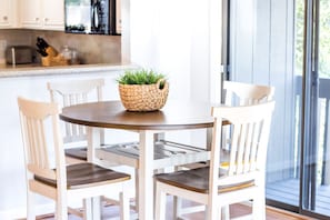 Une table de salle à manger ronde, entourée de quatre chaises blanches, un panier en osier contenant des plantes vertes et, au fond, une cuisine.