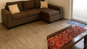 Un canapé d’angle marron avec deux coussins beige, un banc en bois surmonté d’un chemin de table rouge à motifs, et un plancher en bois.