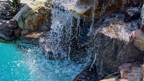 A rock waterfall over a pool with a palm tree nearby. - Elegant 4BR Pool Home + Guest House | Walk to Beach (Naples)